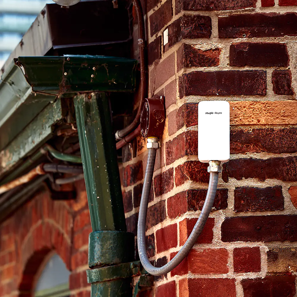 A white rectangular access point installed on a red brick building. The device has a Ruijie Reyee logo on it.