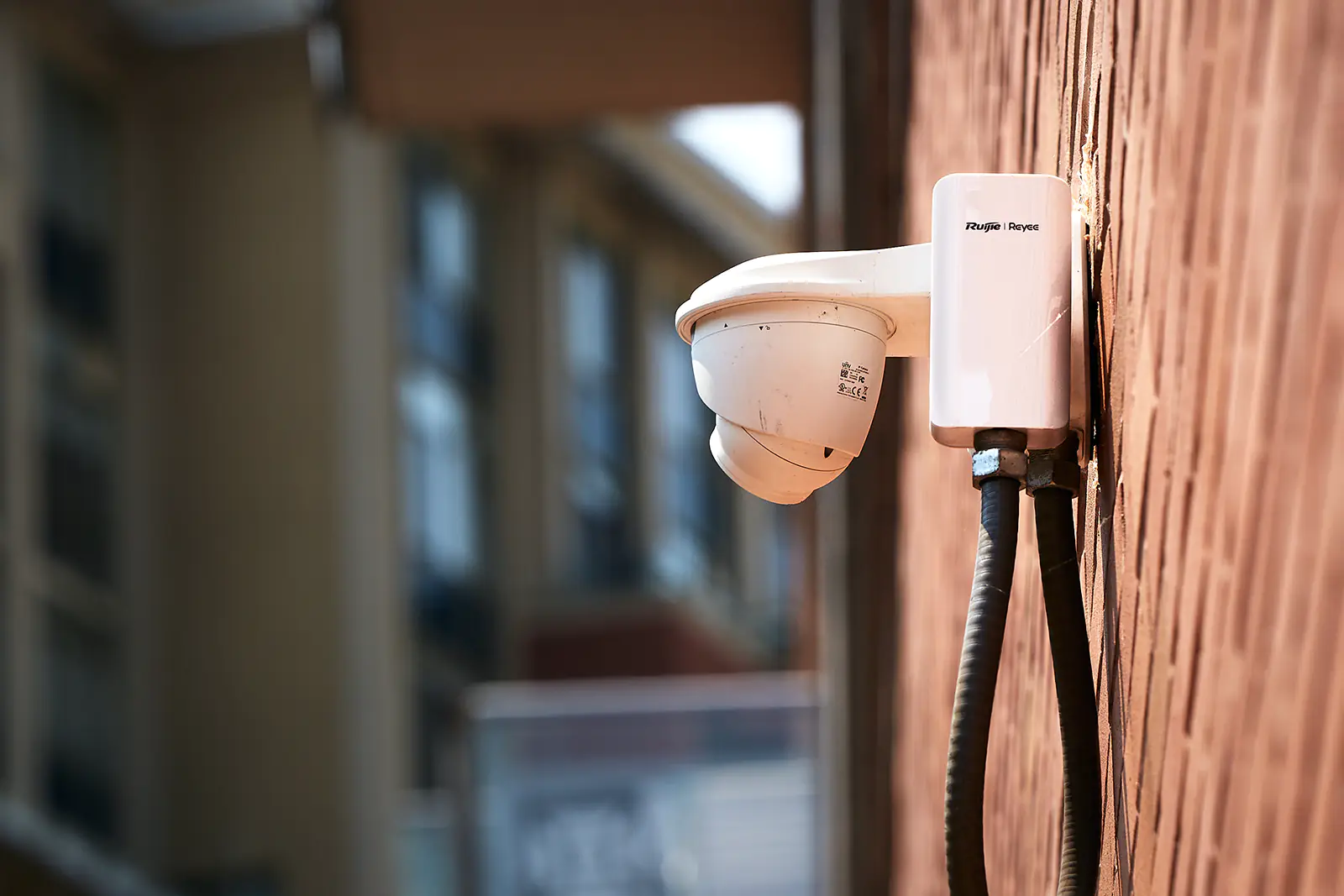 A security camera and a wireless P2P bridge mounted on a brick wall with apartment buildings in the background.